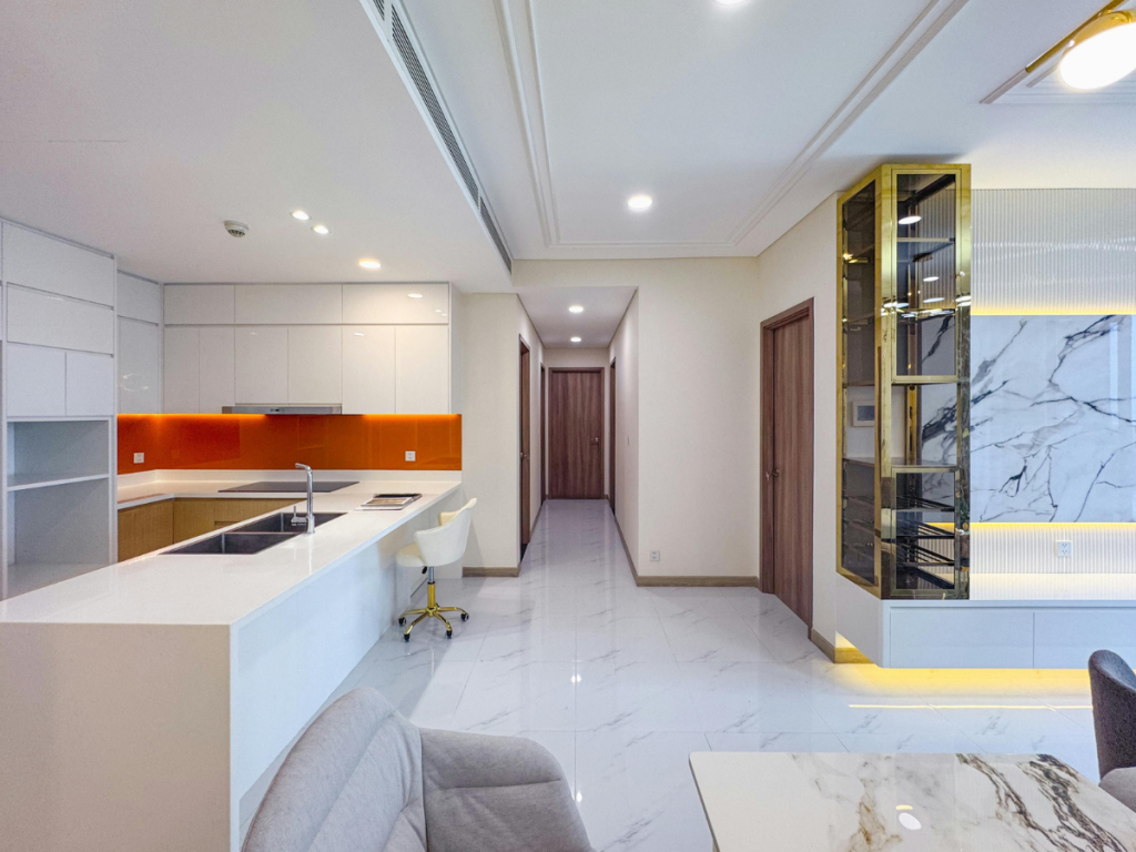 Modernes Apartment-Interieur mit weißer Küche und Wohnzimmer, orangefarbener Küchenrückwand, Marmorboden, eingebauten Regalen und einem Flur, der zu Holztüren führt.