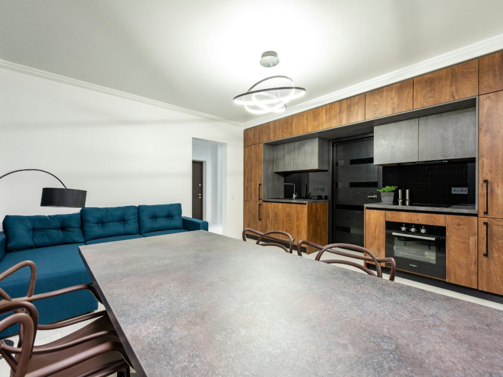 Moderne Küche und Wohnzimmer mit Holzschränken, Einbaugeräten, einem grauen Esstisch, braunen Stühlen und einem blauen Sofa vor weißer Wand.