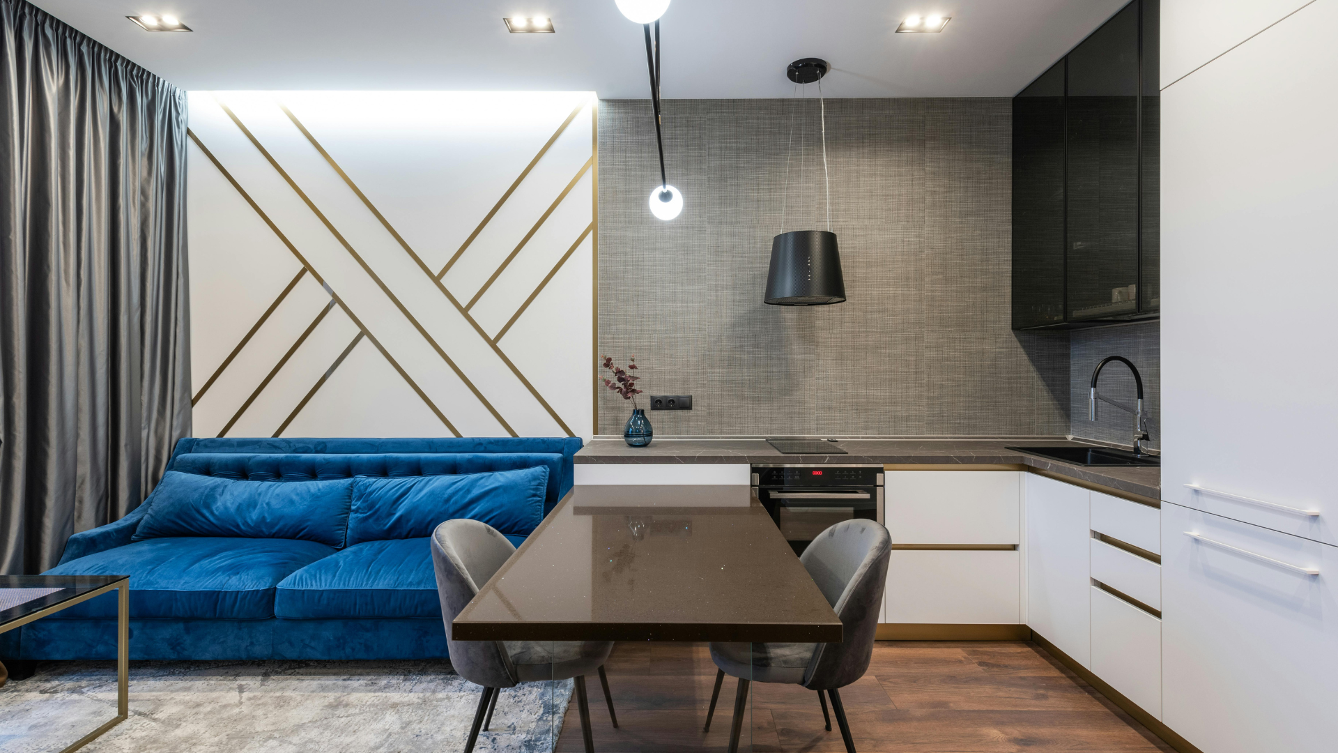 Moderne Küche und Wohnzimmer mit blauem Sofa, geometrischem Wanddesign, schwarzem Esstisch mit Stühlen und eleganten weißen Schränken bei sanfter Beleuchtung.