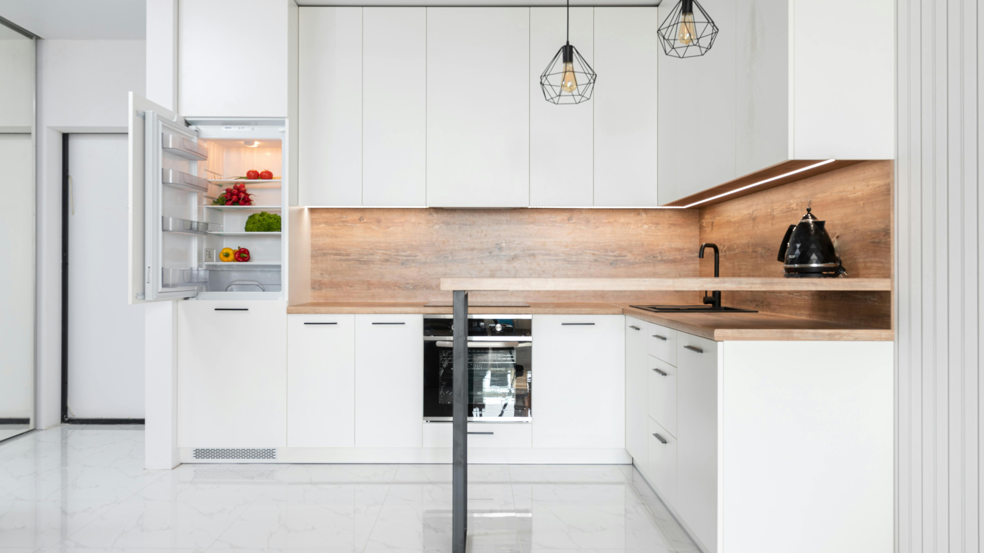 Moderne weiße Küche mit Holzakzenten und offenen Regalen, offenem Kühlschrank mit Lebensmitteln, schwarzem Wasserhahn, eingebautem Backofen und Deckenhängelampen.
