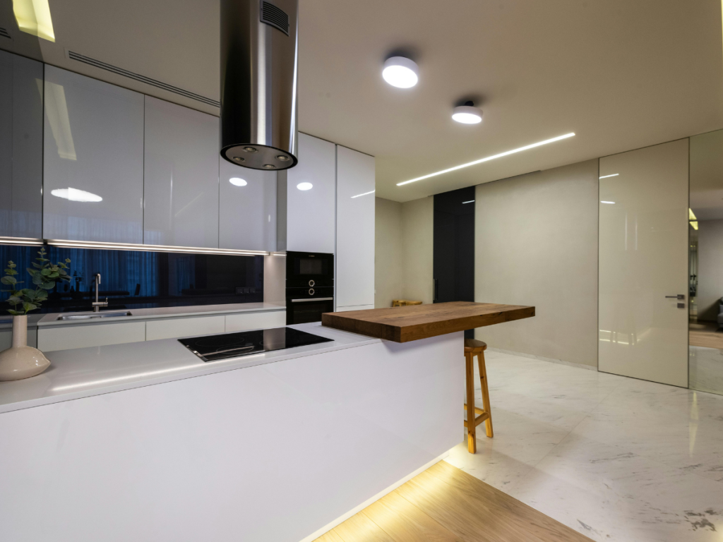 Moderne Küche mit glänzend weißen Schränken, Einbaugeräten und einer Frühstückstheke aus Holz mit Hocker. Dies ist ein Beispiel für den schlanken Minimalismus, der in modernen Küchen zu finden ist. Das Ganze wird von hellen Deckenlampen beleuchtet.
