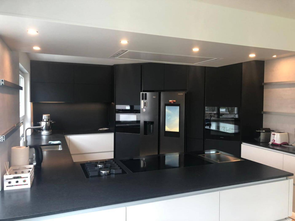 Moderne Küche mit schwarzen Schränken, Einbaugeräten, einem Kühlschrank mit Display und einer schlanken schwarzen Arbeitsplatte mit Deckenstrahlern.