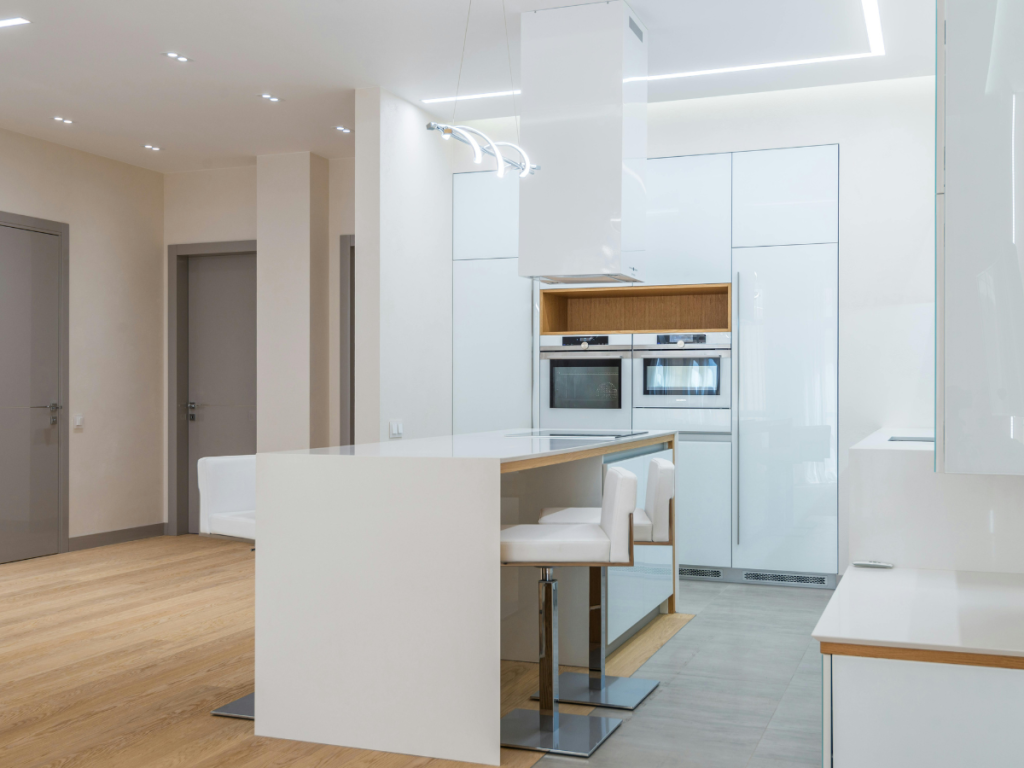 Moderne Küche mit weißen Schränken, Einbauöfen, einer Kücheninsel mit zwei weißen Stühlen, Holzboden und minimalistischer Beleuchtung.