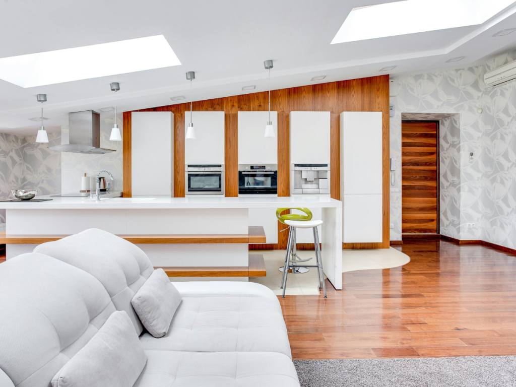 Moderne Küche und Wohnzimmer mit weißen Schränken, Einbaugeräten, einer Kücheninsel mit Barhocker und weißem Sofa im Vordergrund, Holzboden und Tageslicht durch Oberlichter.