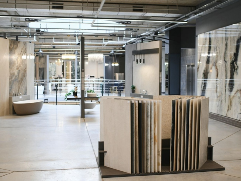 Grote showroom met tentoongestelde tegels en natuurstenen platen in diverse vloerpatronen, moderne badkamerinrichting inclusief ligbad en veel natuurlijk licht via de dakramen.