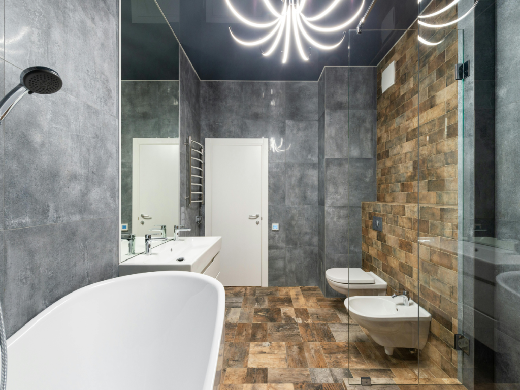 Modernes Badezimmer mit grauen und braun gefliesten Wänden, einer freistehenden weißen Badewanne, einem wandhängenden Waschbecken, einem Bidet, einer Toilette und moderner Beleuchtung mit einem stilvollen Kronleuchter.