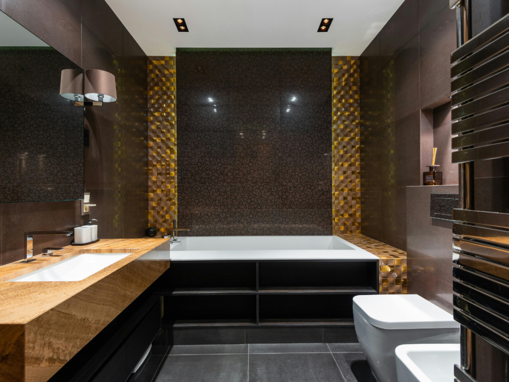 Modernes Badezimmer mit großer Badewanne, Holzarbeitsplatte, Doppelwaschbecken, Wandspiegel, goldfarbenen Akzentfliesen hinter der Badewanne und stilvoller Beleuchtung, die eine warme Atmosphäre schafft.