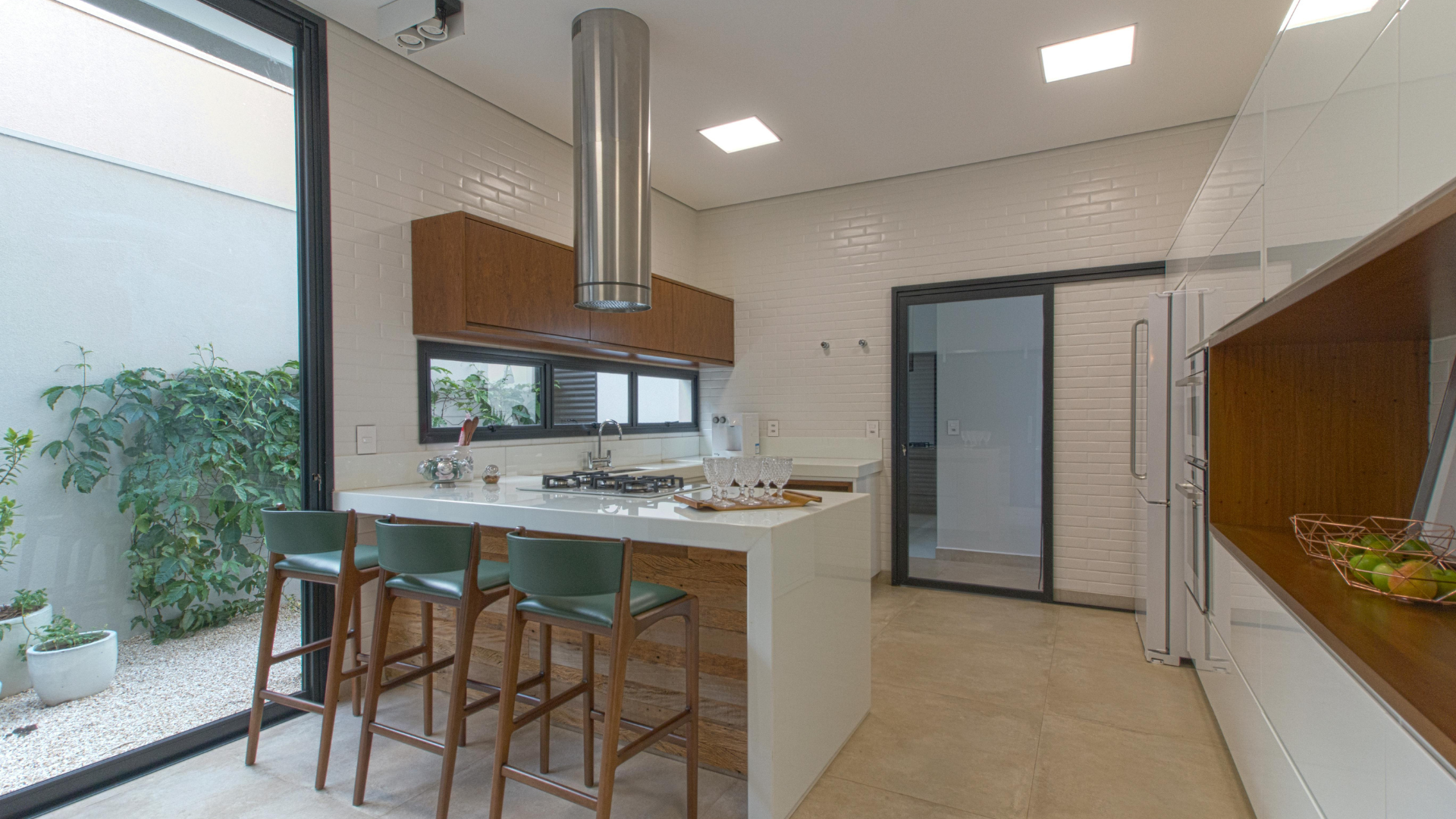 Moderne Küche mit weißen Wänden, großem Fenster, Kücheninsel mit Sitzgelegenheit für drei Personen, Einbaugeräten und Blick auf einen kleinen Garten. Ideal für alle, die moderne Küchen lieben.