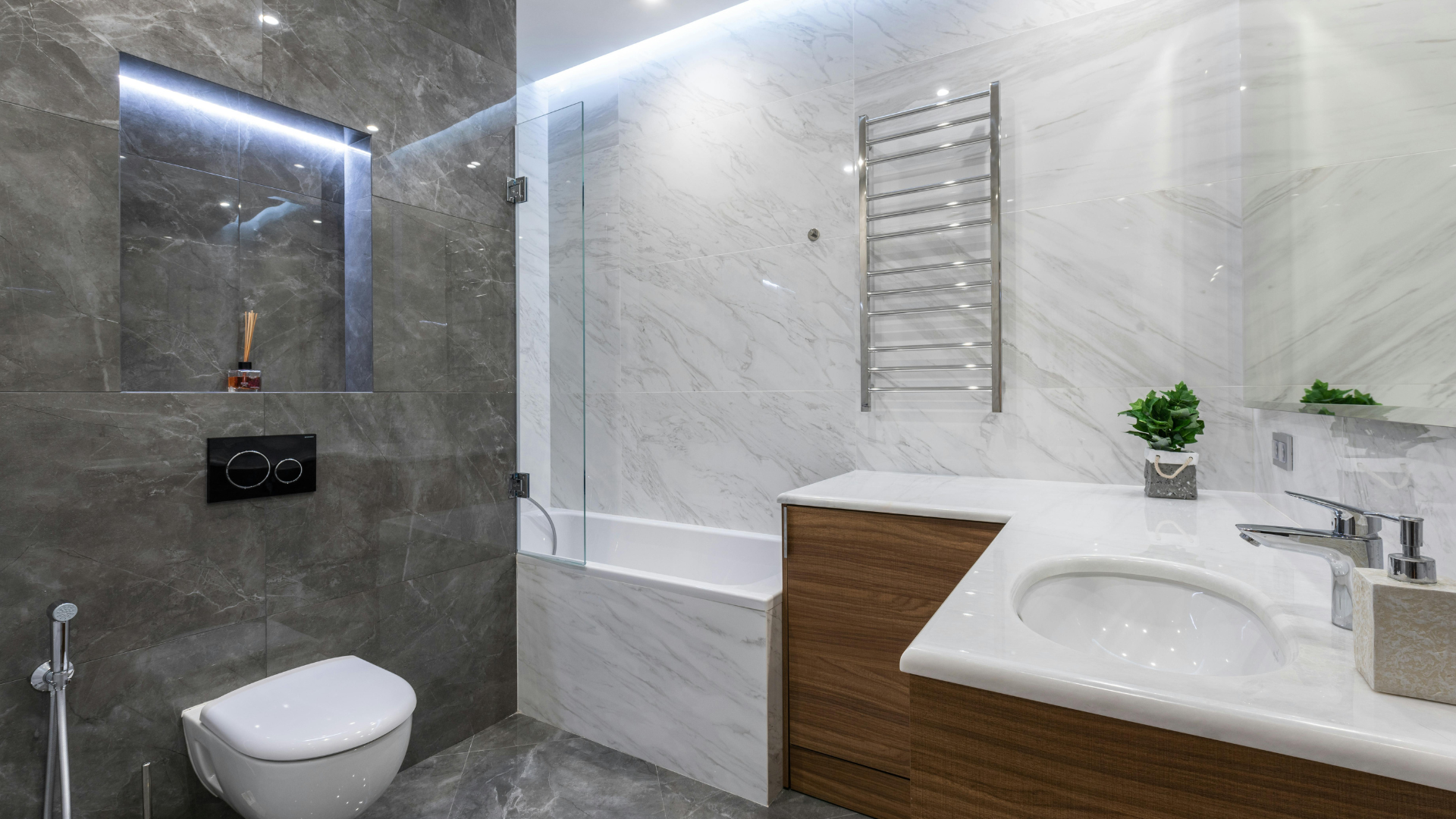 Modernes Badezimmer mit Marmorwänden, Beleuchtung über der Hängetoilette, eingebauter Badewanne, Holzwaschbecken, Handtuchwärmer und einer Topfpflanze auf der Arbeitsplatte.