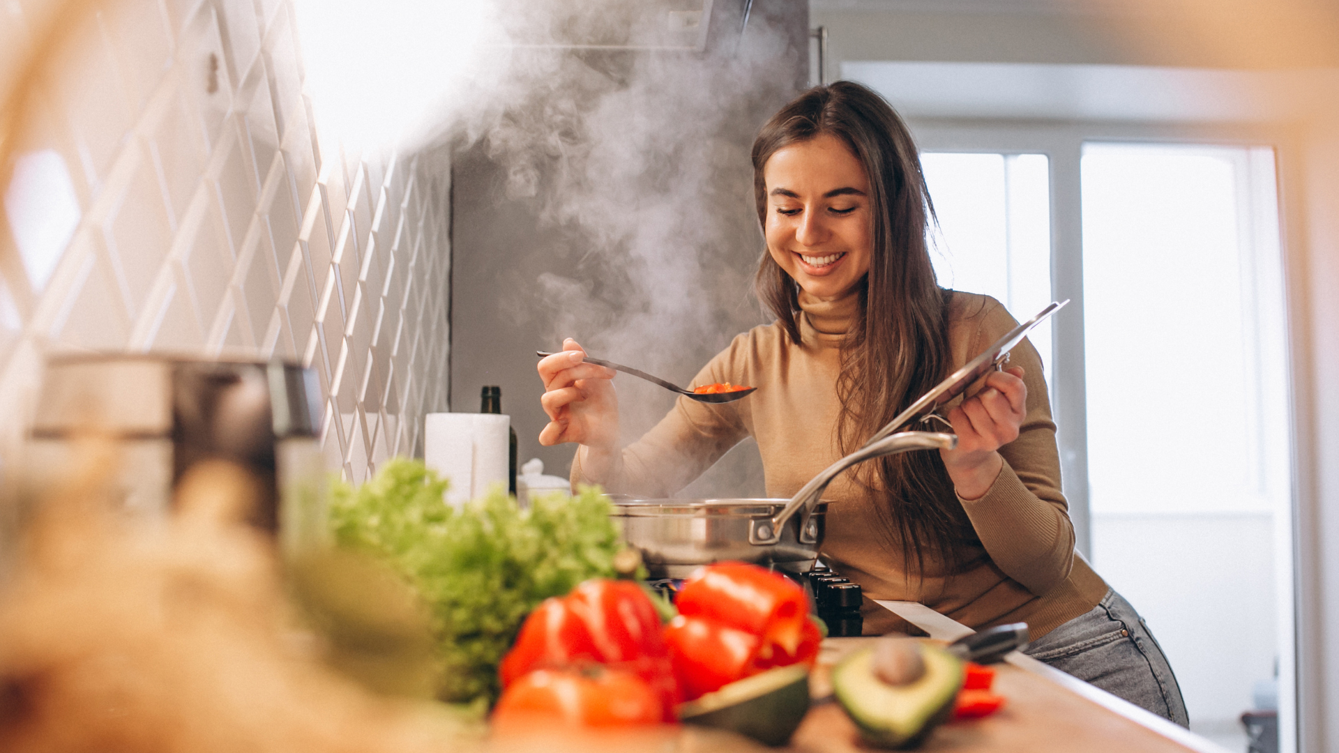Eine Frau in der Küche probiert Suppe aus einer Pfanne auf dem Herd, auf der Theke steht frisches Gemüse wie Paprika und Avocado. Dampf steigt aus der Pfanne auf und komplizierte Bodenmuster verleihen dem gemütlichen Raum Charme.