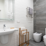 Modernes privates Badezimmer mit weißem Waschbecken, Holzaccessoires, Handtuchhalter, Spiegelschrank, Bidet und WC vor einer strukturierten Wand mit grauen Fliesenakzenten.