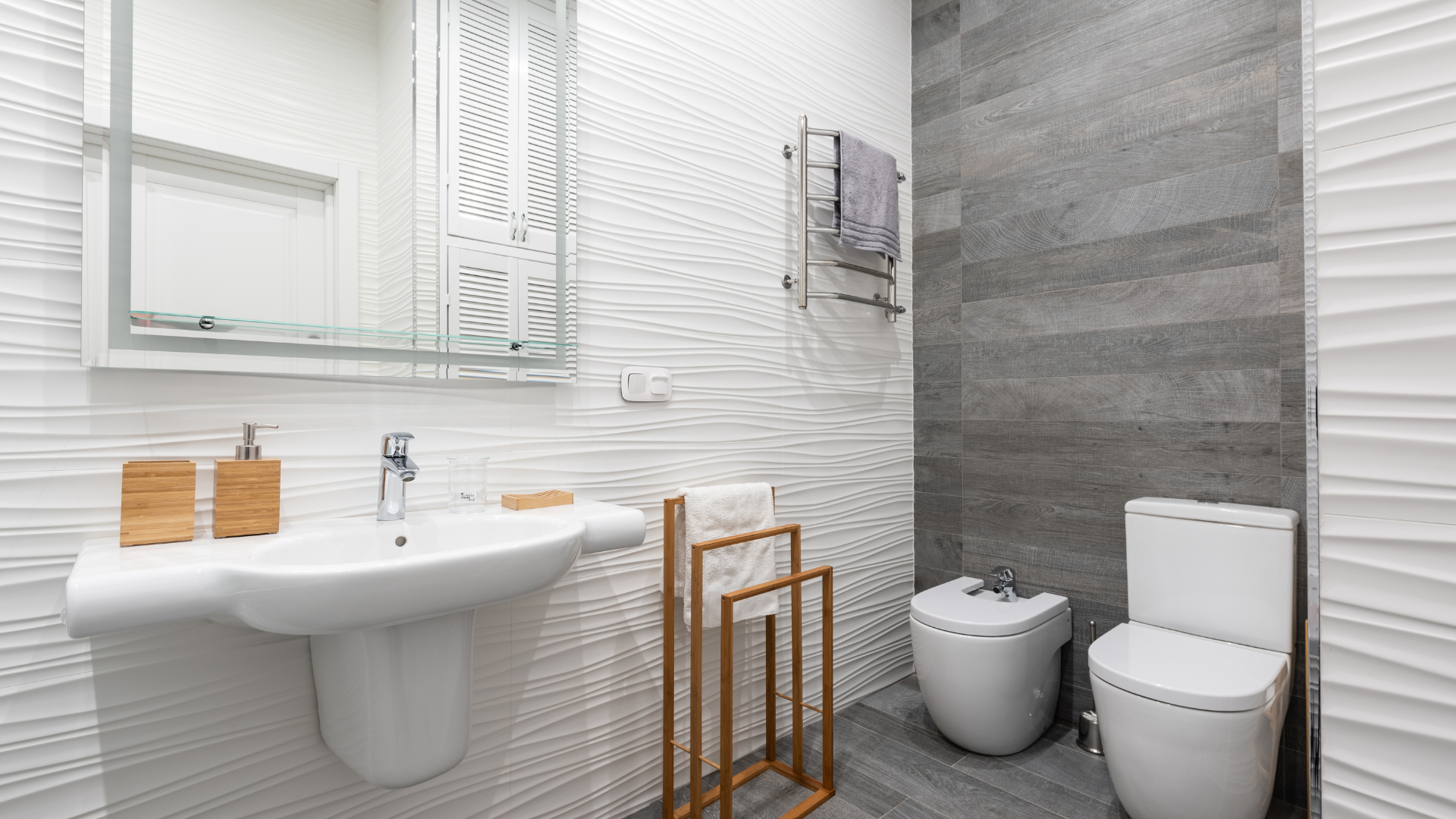 Modernes privates Badezimmer mit weißem Waschbecken, Holzaccessoires, Handtuchhalter, Spiegelschrank, Bidet und WC vor einer strukturierten Wand mit grauen Fliesenakzenten.
