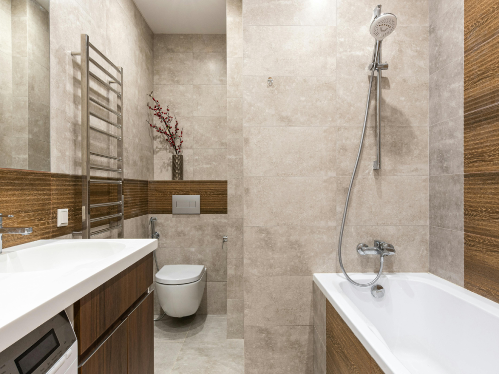 Modernes privates Badezimmer mit beigen Fliesen, Wandtoilette, Badewanne mit Duschkopf, beheiztem Handtuchhalter und einem Waschtischunterschrank mit Waschbecken und Blumenvase.