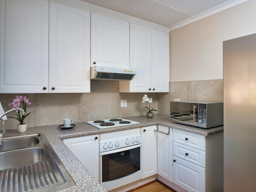 Moderne, kleine Küchen mit weißen Schränken, Elektroherd, Backofen, Mikrowelle, Edelstahlspüle und zwei Orchideen in Töpfen auf der Theke.