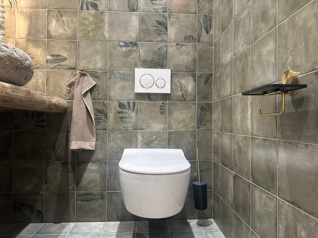 Ein modernes Badezimmer mit grün gemusterten Fliesen, einer Wandtoilette, einem beigen Handtuch an einem Haken, einer Toilettenbürste und einem leeren Toilettenpapierhalter.