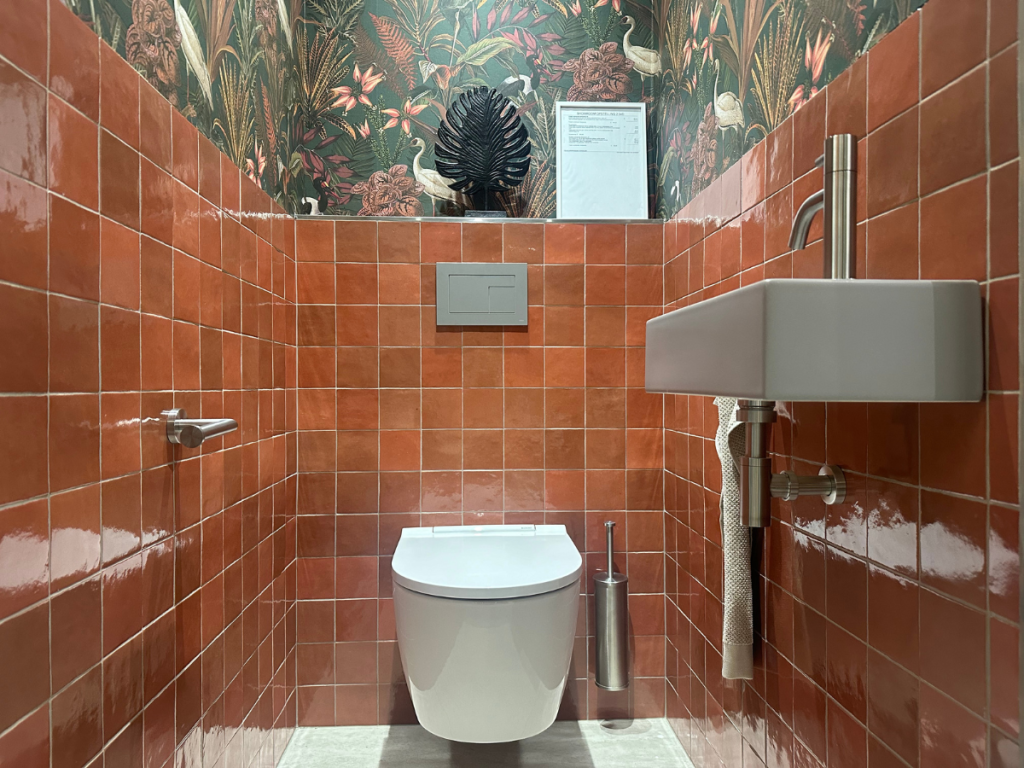 Ein kleines Badezimmer mit orangefarbenen Fliesenwänden, einer wandhängenden Toilette, einem kleinen rechteckigen Waschbecken, einer Toilettenpapierrolle und tropischen Tapeten über den Fliesen.