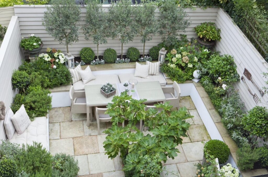 A small, landscaped patio garden, enclosed, including a dining table set surrounded by lush vegetation, including shrubs, potted plants and small trees. This landscaped space has a light stone floor and is surrounded by wooden fences. The landscaping company has designed this landscaping service to create a soothing and harmonious atmosphere.