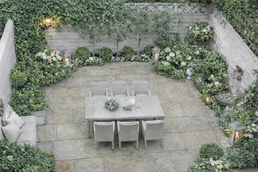 Небольшой ландшафтный сад во внутреннем дворике, огороженный, включая обеденный стол, окруженный пышной растительностью, включая кустарники, растения в горшках и небольшие деревья. Это благоустроенное пространство имеет светлый каменный пол и окружено деревянными заборами. Компания по ландшафтному дизайну разработала эту услугу по ландшафтному дизайну для создания успокаивающей и гармоничной атмосферы.