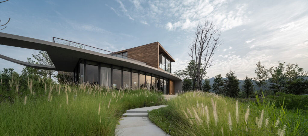 This project unfolds around a contemporary architect's villa, with large windows and a wooden upper section, integrated into an environment where architecture meets nature. The surroundings are designed with tall grass and trees to create a natural and harmonious setting. A concrete path with curved lines guides visitors to the main entrance, combining aesthetics and functionality.