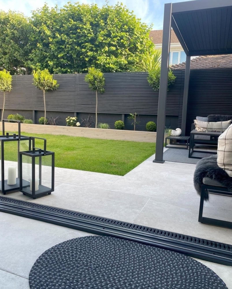 On a modern terrace with a stylish pergola, cushioned seating and decorative lanterns add charm. In the background, a landscaped garden features a manicured lawn and small plants, framed by a black slatted fence and tall hedges. These developments are typical of a landscaping service carried out by a landscaping company or an experienced landscaping company. The art of landscaping is reflected in every detail of this harmonious outdoor space.