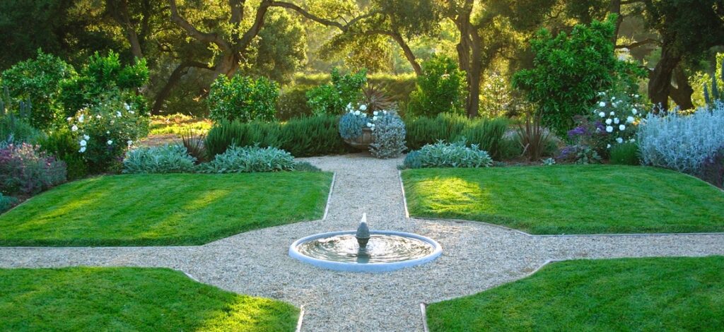 A symmetrical landscaped garden with a central circular fountain, gravel walkways dividing the green lawns into four sections, surrounded by various plants and trees in the background. This type of design can be the result of the work of a landscaper or a landscaping company that specializes in creating and maintaining gardens. A landscaping company could offer a complete landscaping service to create such a harmonious and well-structured space.