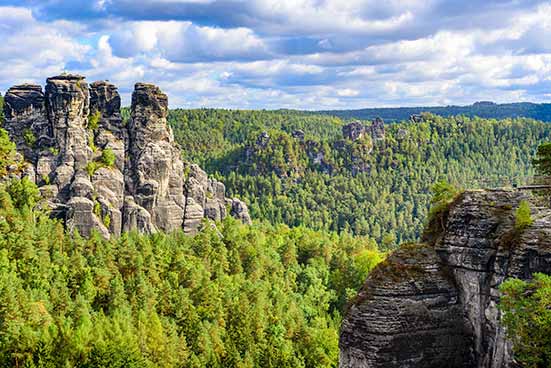 Under a cloudy sky, rock formations rise above a dense forest, revealing a natural landscape of lush greenery and rugged cliffs.