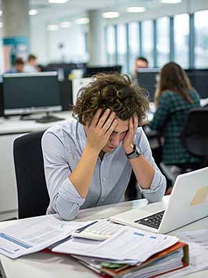 A person with curly hair sits at a desk in front of a laptop and holds his head with both hands. Papers and a pen are scattered on the desk. An office environment can be seen in the background.