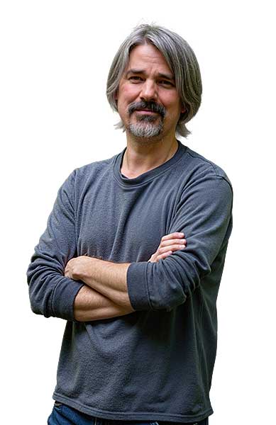 A man with gray hair and a beard stands with his arms crossed in a long-sleeved gray shirt and jeans in front of a white background.