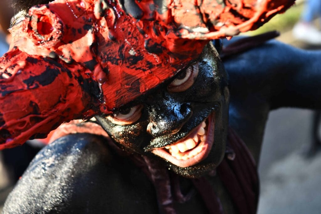 Person with face paint smiling, wearing a large red and black headdress, embodying the spirit of the Diyandi Festival.