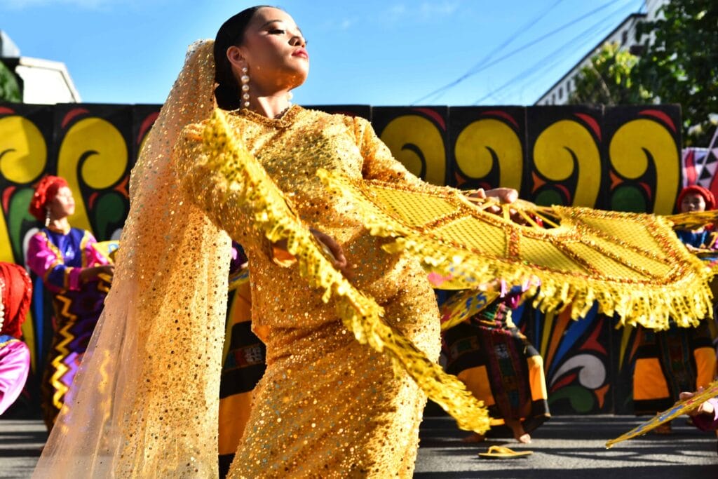 A woman in a sparkling gold outfit dances with a fan at the vibrant Diyandi Festival.
