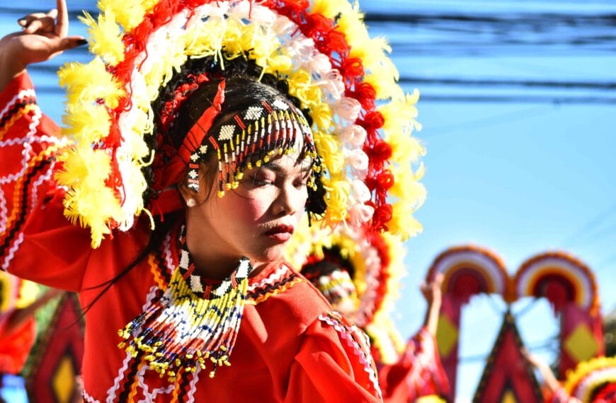Mananayaw sa pulang costume, makulay na headdress, mata sarado, pagdiriwang Diyandi Festival laban sa isang maliwanag na asul na kalangitan.