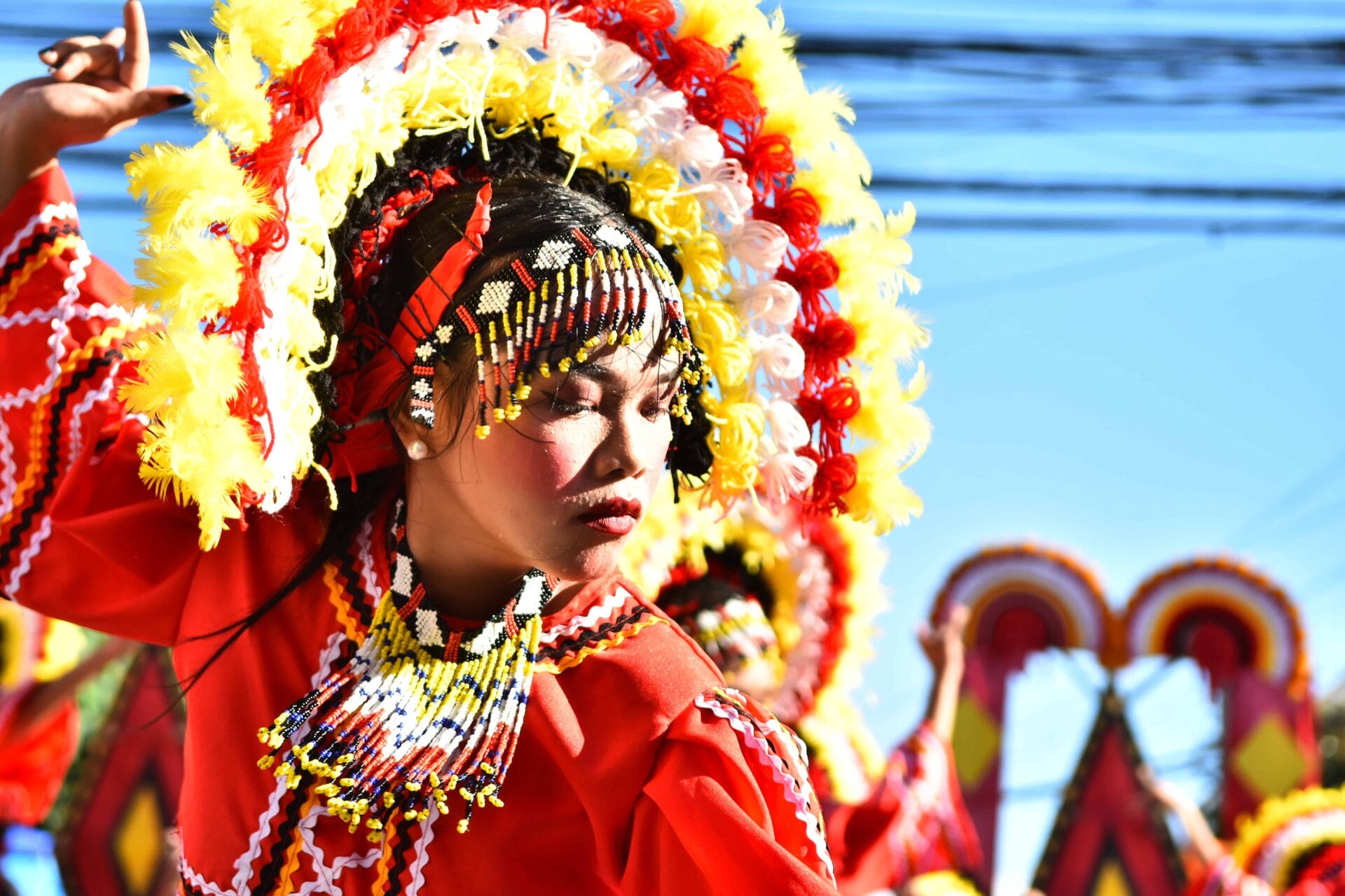 Mananayaw sa pulang costume, makulay na headdress, mata sarado, pagdiriwang Diyandi Festival laban sa isang maliwanag na asul na kalangitan.