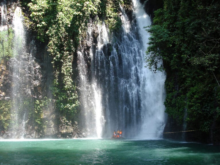 Kagila-gilalas na Tinago Falls cascading sa isang turkesa pool, malago berde enveloping; mga taong naka orange vests sa ibaba.