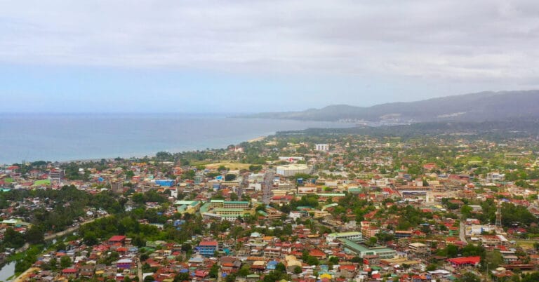 Aerial view ng isang coastal Iligan City na may makukulay na rooftops, mayabong na luntian, at burol sa ilalim ng maulap na kalangitan.