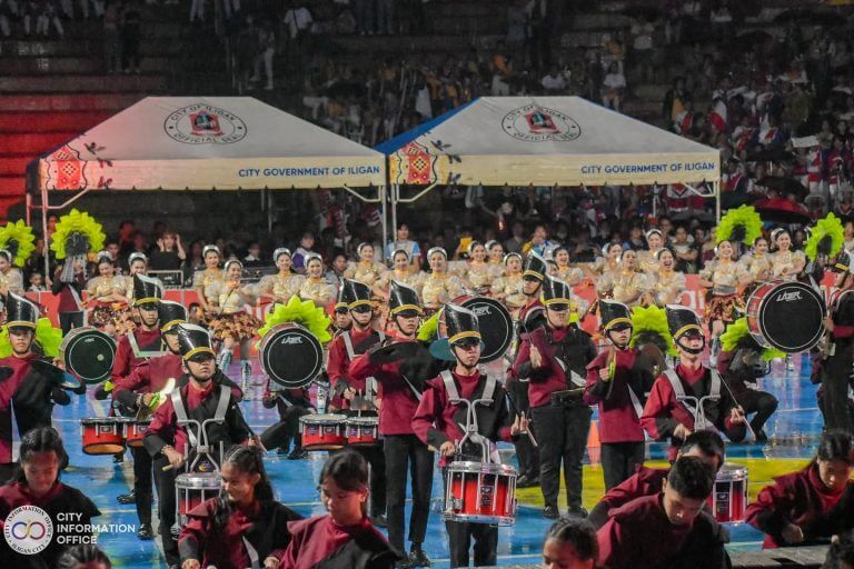 Isang marching band na nakasuot ng maroon ang tumutugtog sa Drum and Lire Fest, na may mga mananayaw at tolda sa Iligan sa likuran nila.
