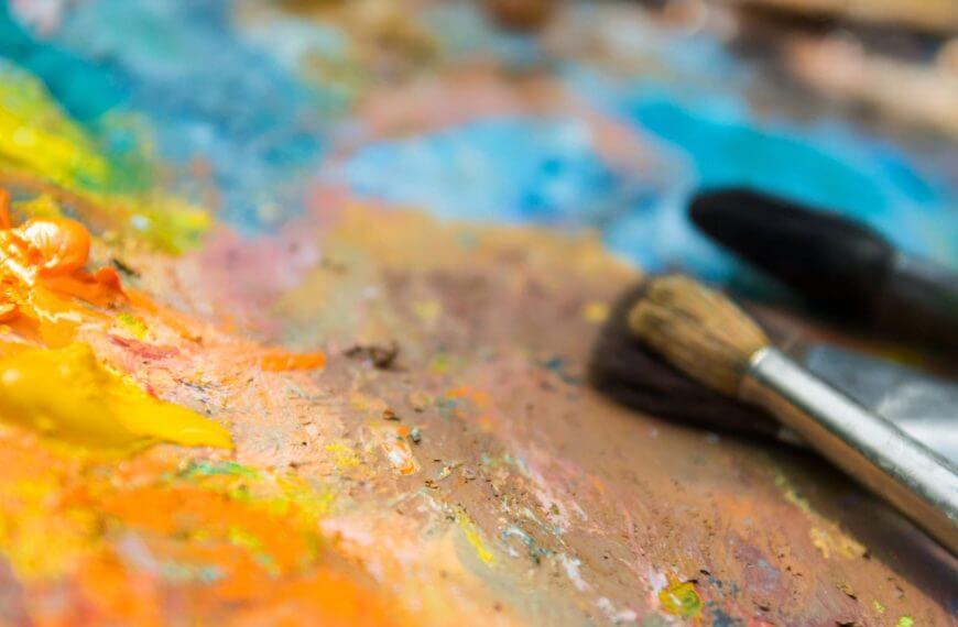 Tatlong paintbrushes ang nakasalalay sa magulo na palette sa Mindanao Art, na natatakpan ng matingkad na dilaw, orange, at asul na pintura.