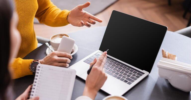 Tinalakay ng dalawang tao ang mga pagkakataon sa freelancing sa isang mesa na may laptop, kape, at telepono sa isang maginhawang setting.