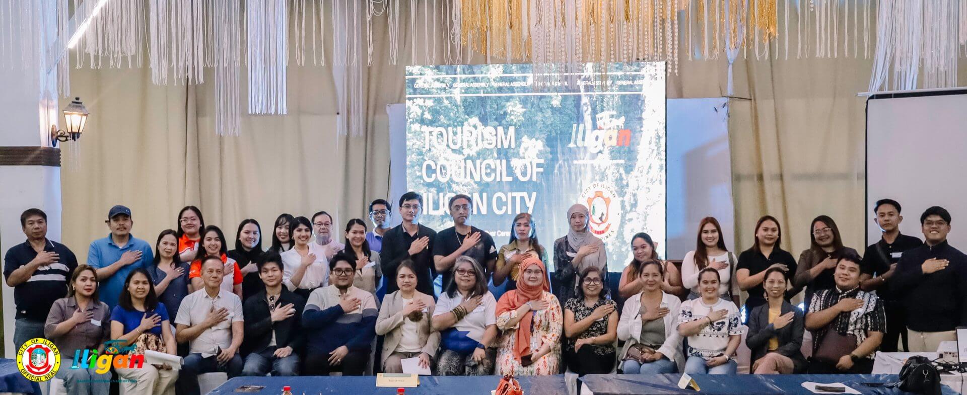a large group poses indoors, smiling with hands over hearts, in front of a tourism council presentation screen.