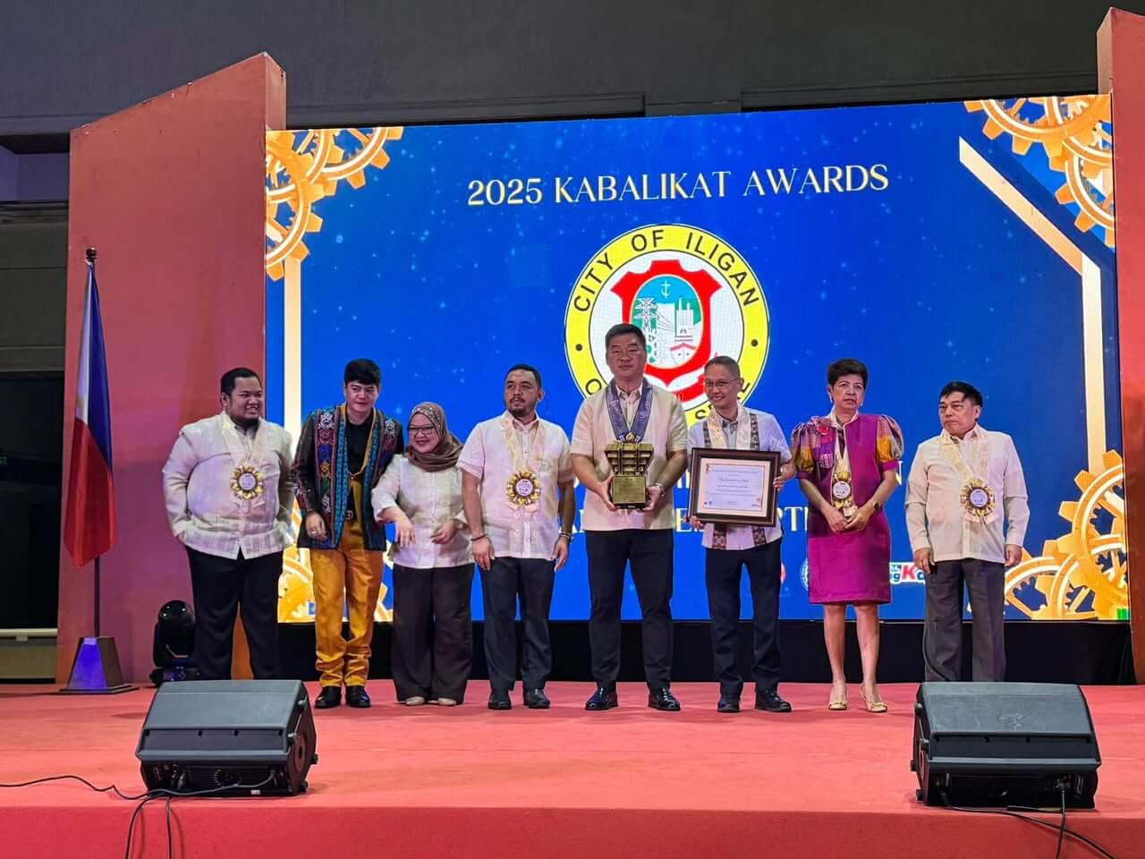 eight people in formal attire pose onstage with tesda kabalikat award trophies at iligan’s 2025 ceremony.