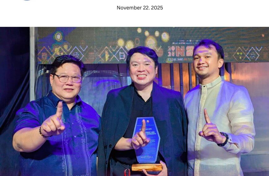 three men smile and hold up one finger; the man in the middle holds a blue and gold award on stage.