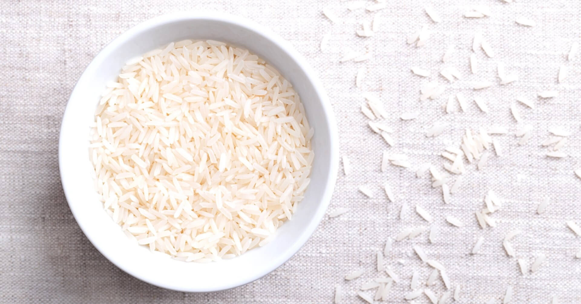 white bowl filled with uncooked dinorado mabango rice, grains scattered on a light fabric.