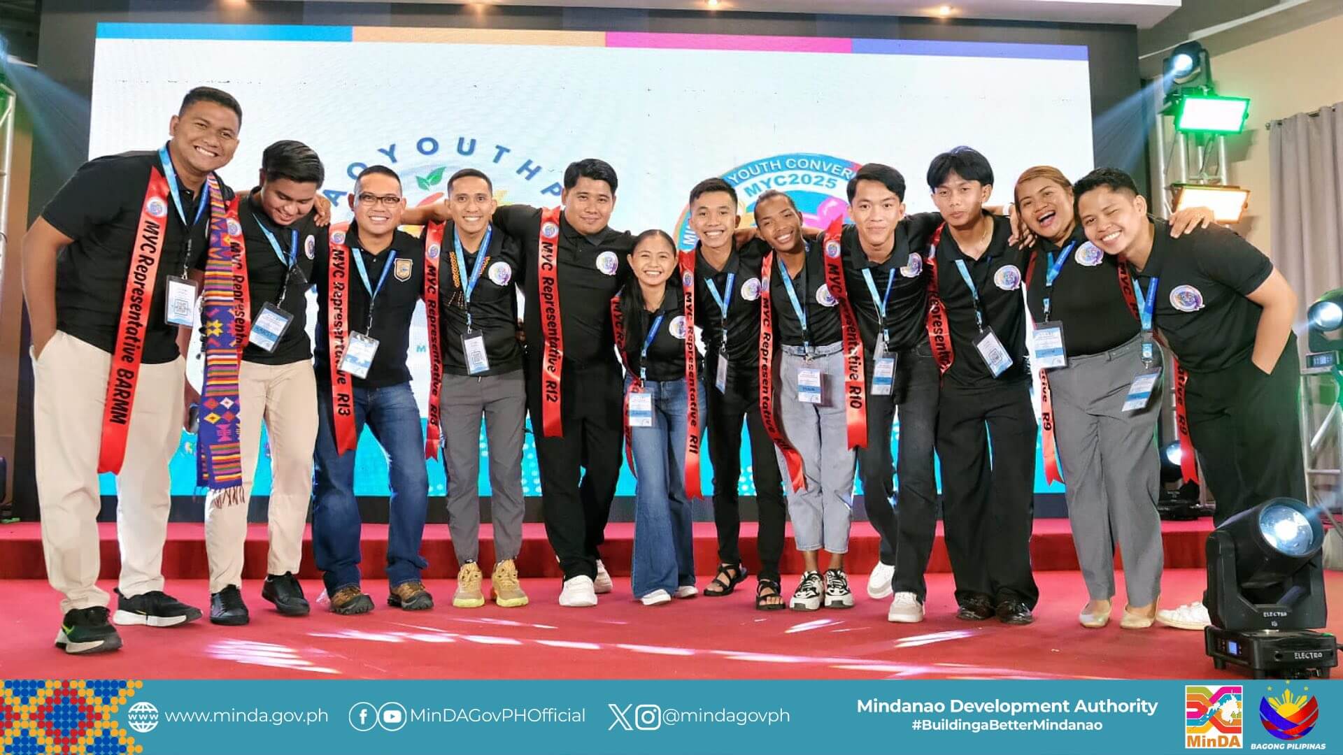 eleven smiling young adults in black shirts pose together on stage at a mindanao youth event.