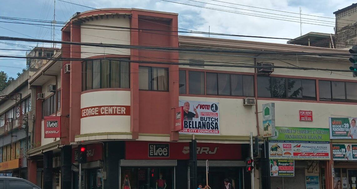 a two story commercial real estate building with shops, signs, and a political campaign banner on the corner.
