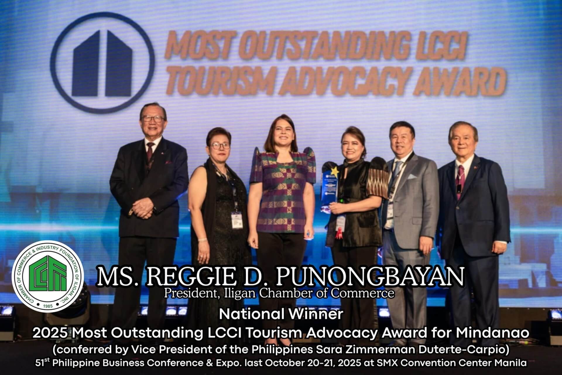 six people stand on stage holding an award, with a blue backdrop and text about a tourism advocacy award.