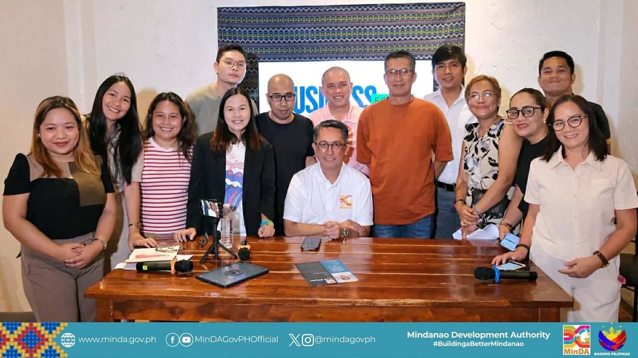 fifteen people smiling, posing together indoors behind a wooden table with microphones and papers.