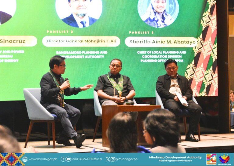 three panelists sit on stage discussing at an event, with colorful banners and a logo in the background.