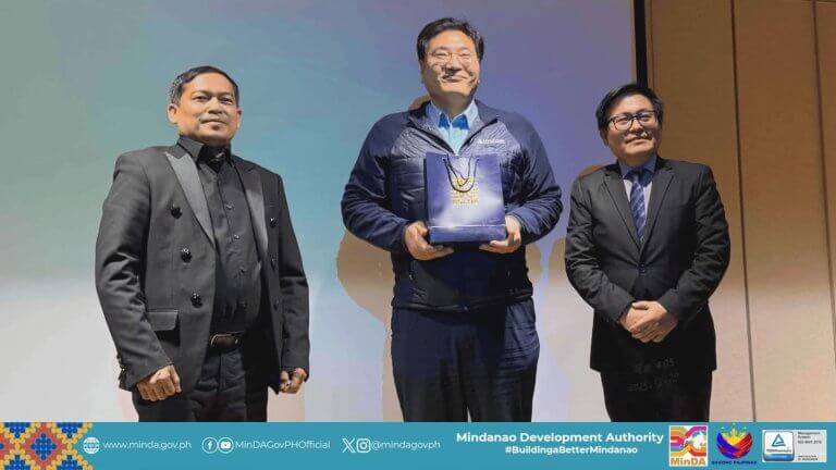 three men in suits stand onstage; the center man with a hyundai gift bag smiles at the camera.