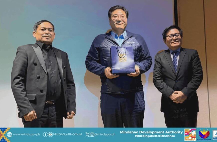 three men in suits stand onstage; the center man with a hyundai gift bag smiles at the camera.