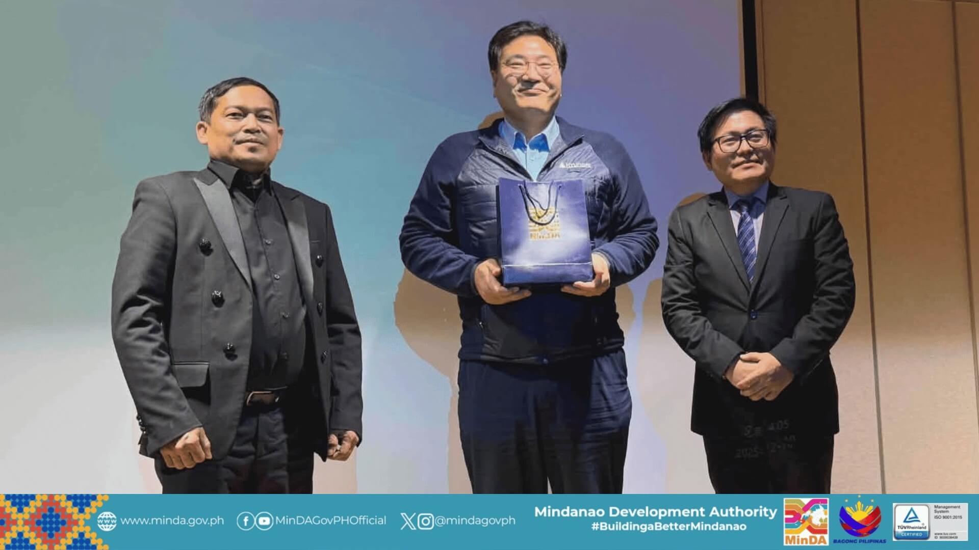 three men in suits stand onstage; the center man with a hyundai gift bag smiles at the camera.