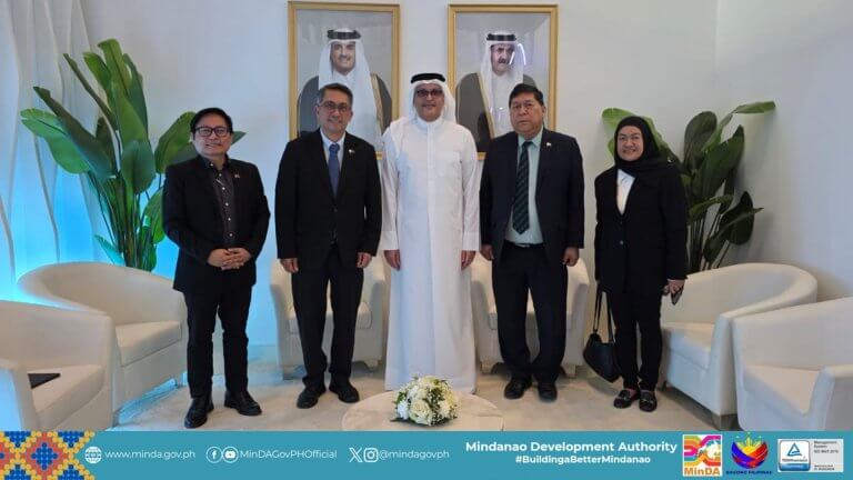 five people in formal attire pose indoors; framed portraits and flags decorate the background.